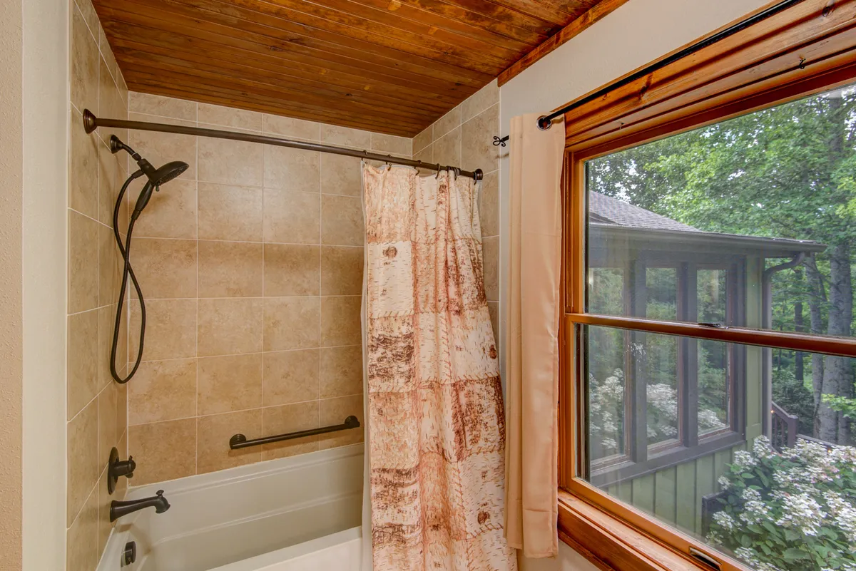 A serene bathroom with a window view, deep soaking bathtub, and a tiled shower with showerhead. Natural light enters the room, creating a tranquil, spa-like atmosphere ideal for unwinding after a day of adventure.