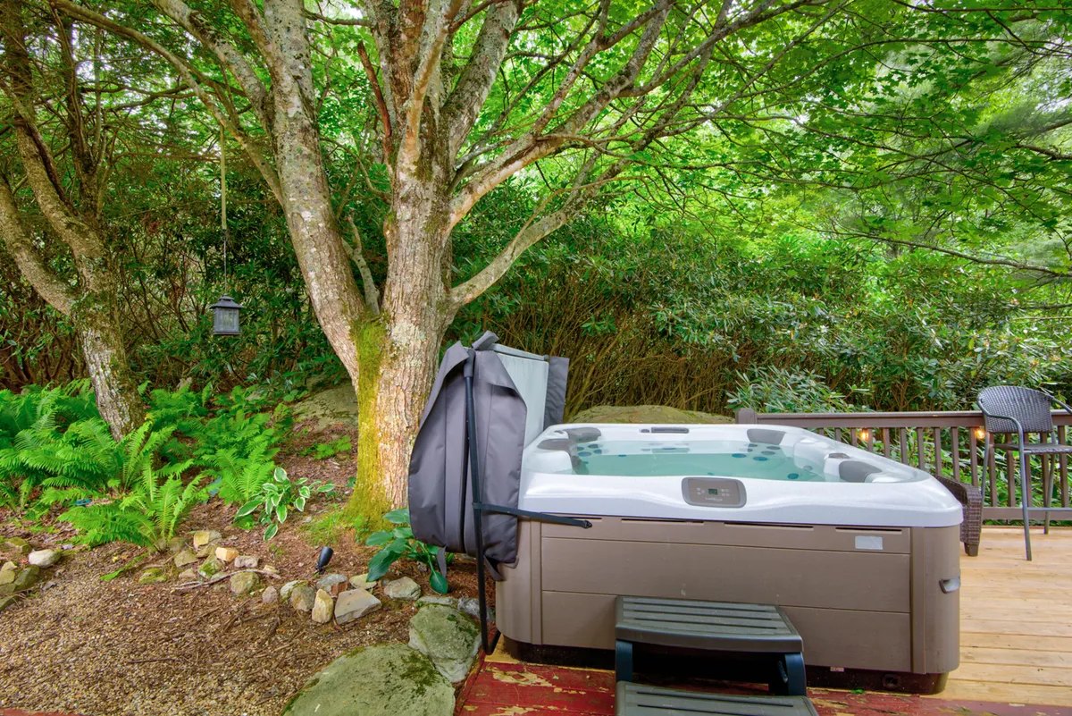 A bubbling hot tub sits nestled among lush greenery and mature trees, creating a secluded oasis for relaxation. The natural setting emphasizes the sensory experience of soaking outdoors, with hints of the deck in the background.