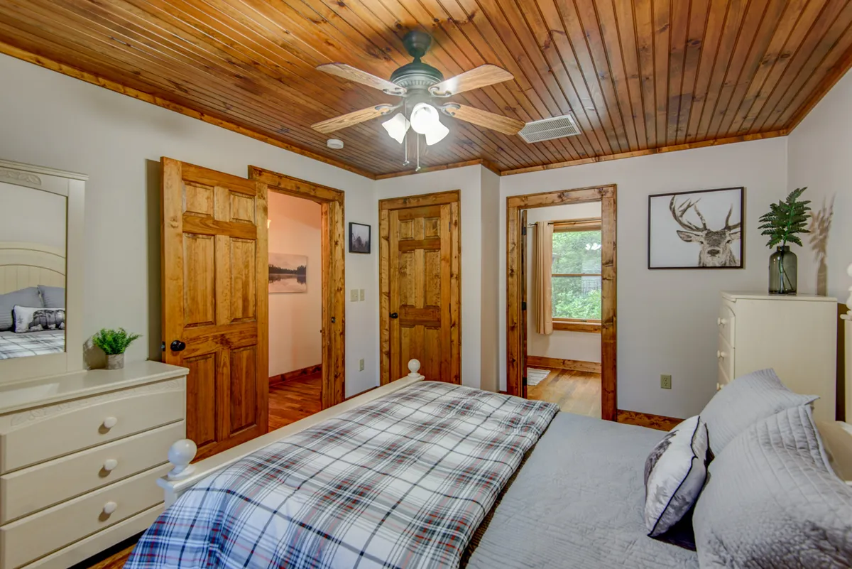 A comfortable bedroom with a plaid bedspread, wooden doors, ceiling fan, and a dresser with a mirror. Decorative wall art of a deer adds a rustic, cozy touch. The room feels warm and inviting, ideal for guests or teens.