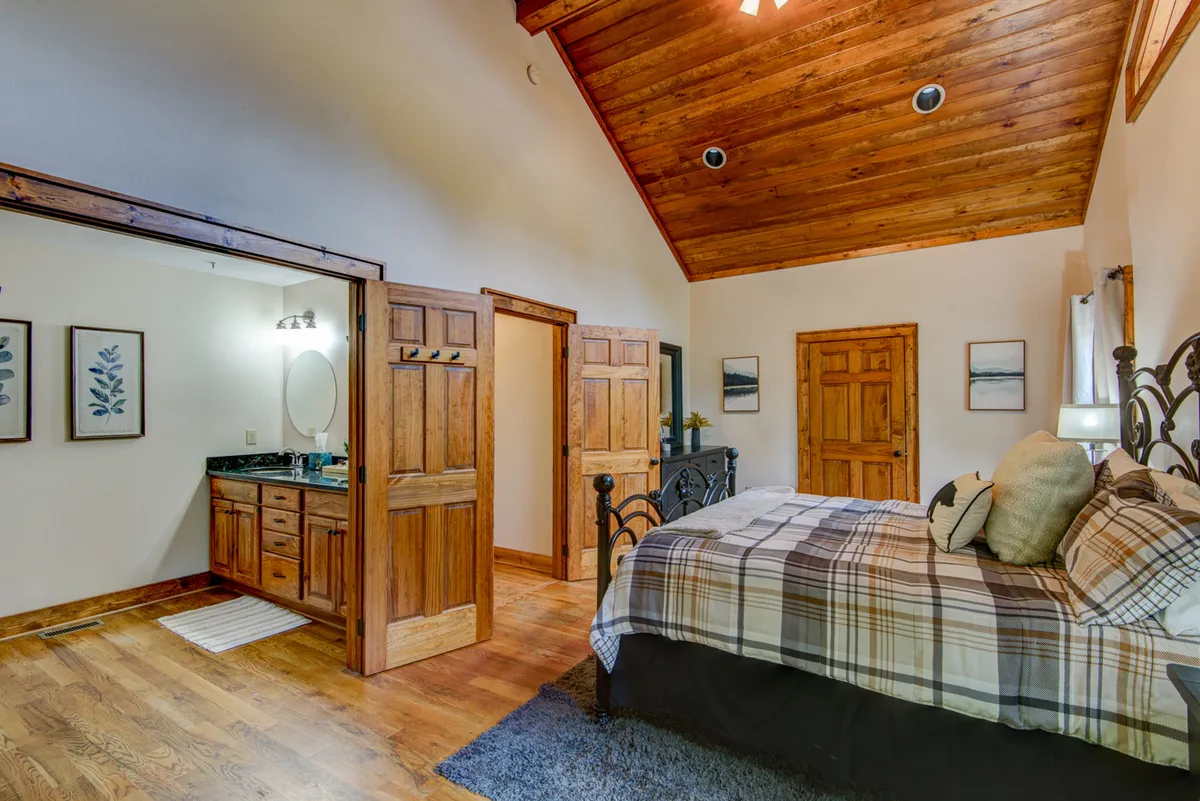 A tranquil bedroom featuring a plush bed topped with plaid bedding beneath a soaring wooden ceiling. The room is bathed in natural light streaming through the windows, and the entrance to an en-suite bathroom is visible, along with tasteful decorative wall art.