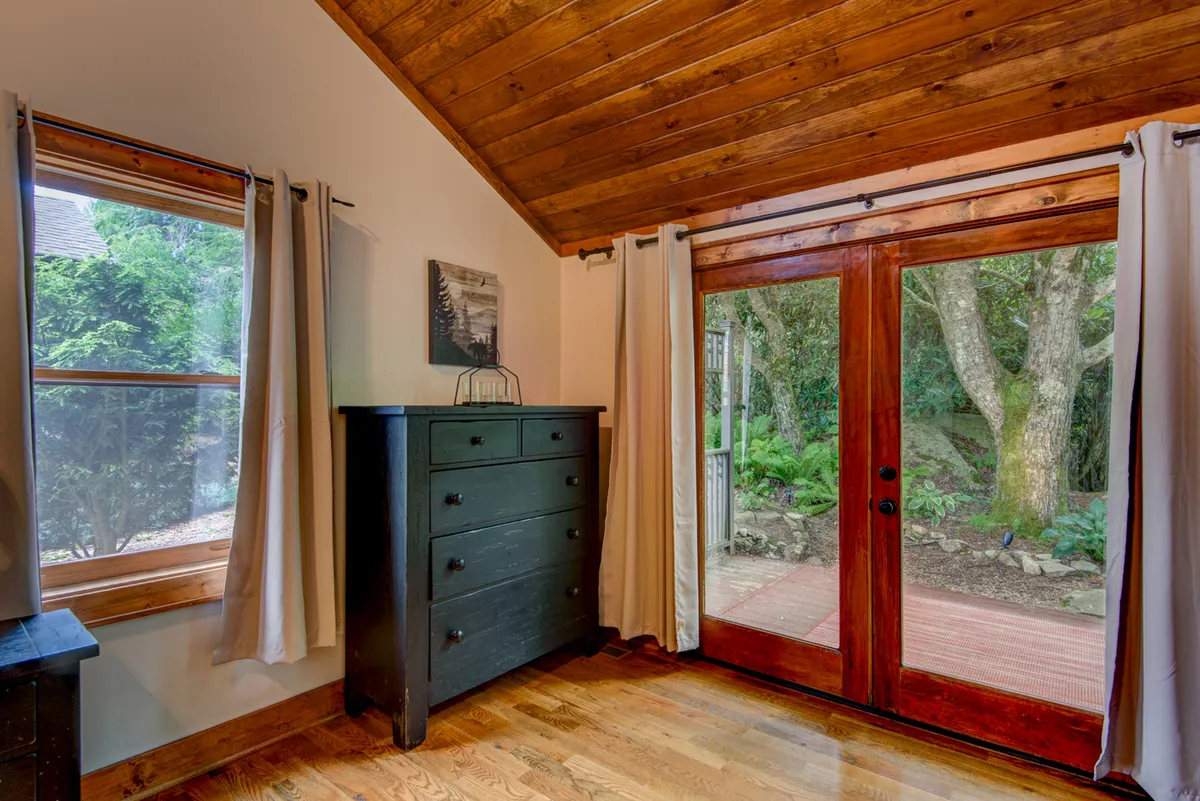 A cozy interior scene featuring wooden elements, a dresser, double doors opening to the outdoors, a window with curtains, and an art piece. The space feels tranquil, warm, and offers a glimpse of the outside, suggesting an easy flow between indoor relaxation and outdoor enjoyment.