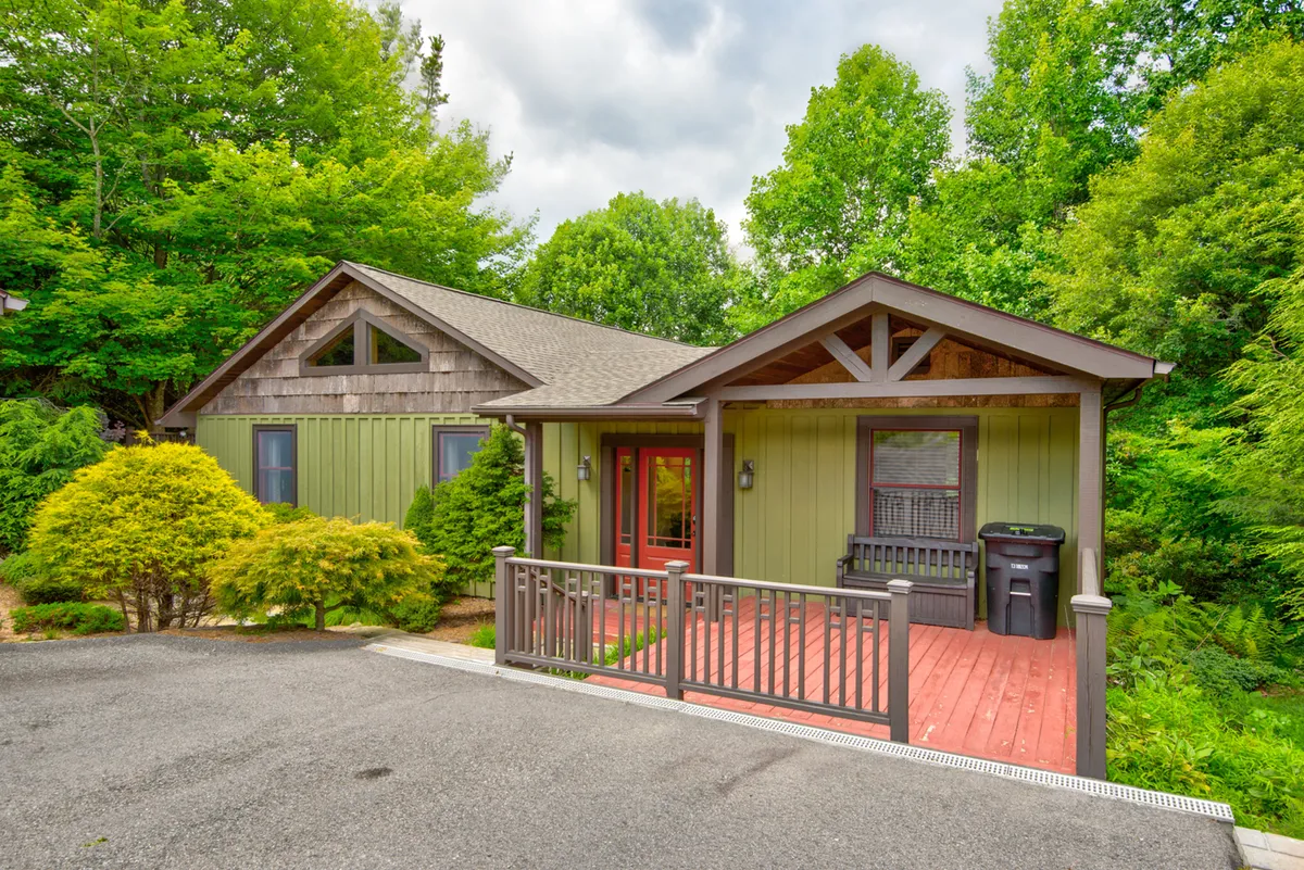 Captured from a slightly different angle, this image showcases the home's green exterior walls, wooden porch railing, and inviting red front door, surrounded by lush greenery. The porch is ready for morning coffee or evening relaxation, while the yard is visible and appears secure and spacious—perfect for both guests and pets.