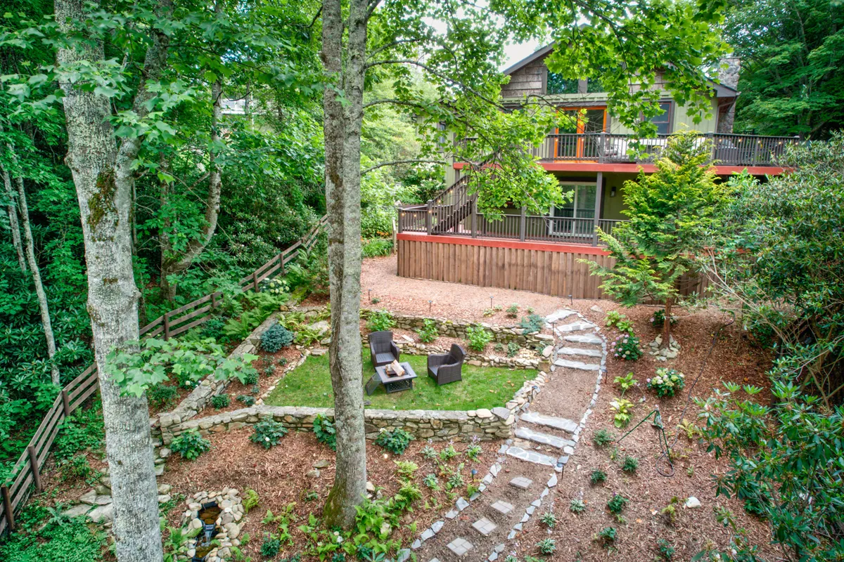 This image showcases the inviting exterior of Bigfoot Bungalow, nestled among lush rhododendron with a charming stone pathway leading to the house. The foreground features a cozy firepit seating area, perfect for evening gatherings, while the wooden deck provides an ideal spot to relax and take in the serene natural surroundings.