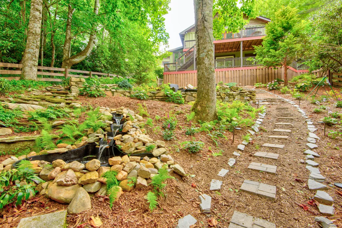 A tranquil exterior scene featuring a stone pathway winding through lush greenery, a water feature, and a deck area attached to the house. The open space invites movement and outdoor activities, and the overall atmosphere is peaceful and welcoming.