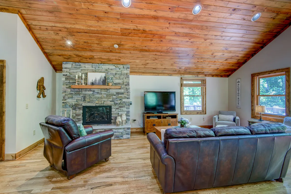 This image features a welcoming living room with leather sofas arranged around a striking stone fireplace. The vaulted wooden ceiling and large windows flood the space with natural light, creating a warm and airy ambiance. A TV is positioned above the fireplace, making this space perfect for relaxation and entertainment.