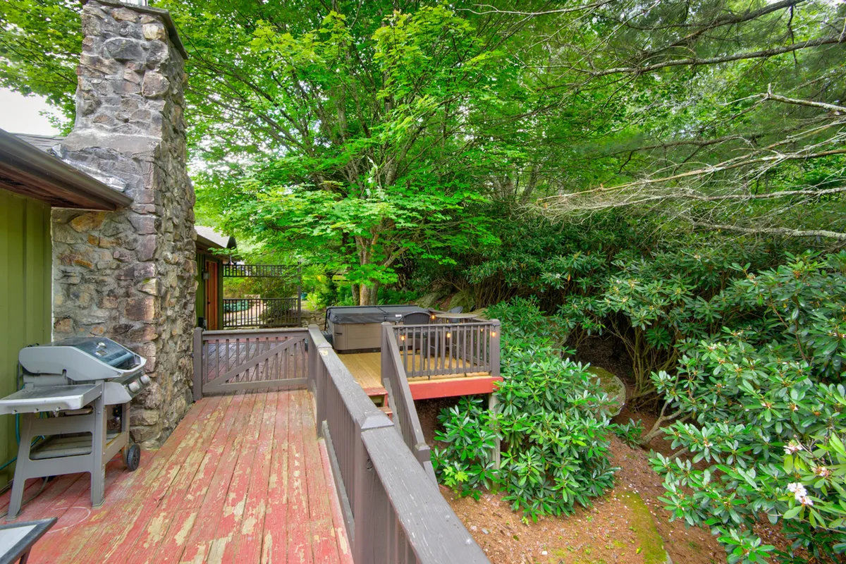 A view of the wooden deck showcases a bubbling hot tub, a classic stone chimney, and lush green surroundings. The deck offers ample space for outdoor dining, barbecuing, and relaxing, all set against the backdrop of dense trees and mountain serenity.