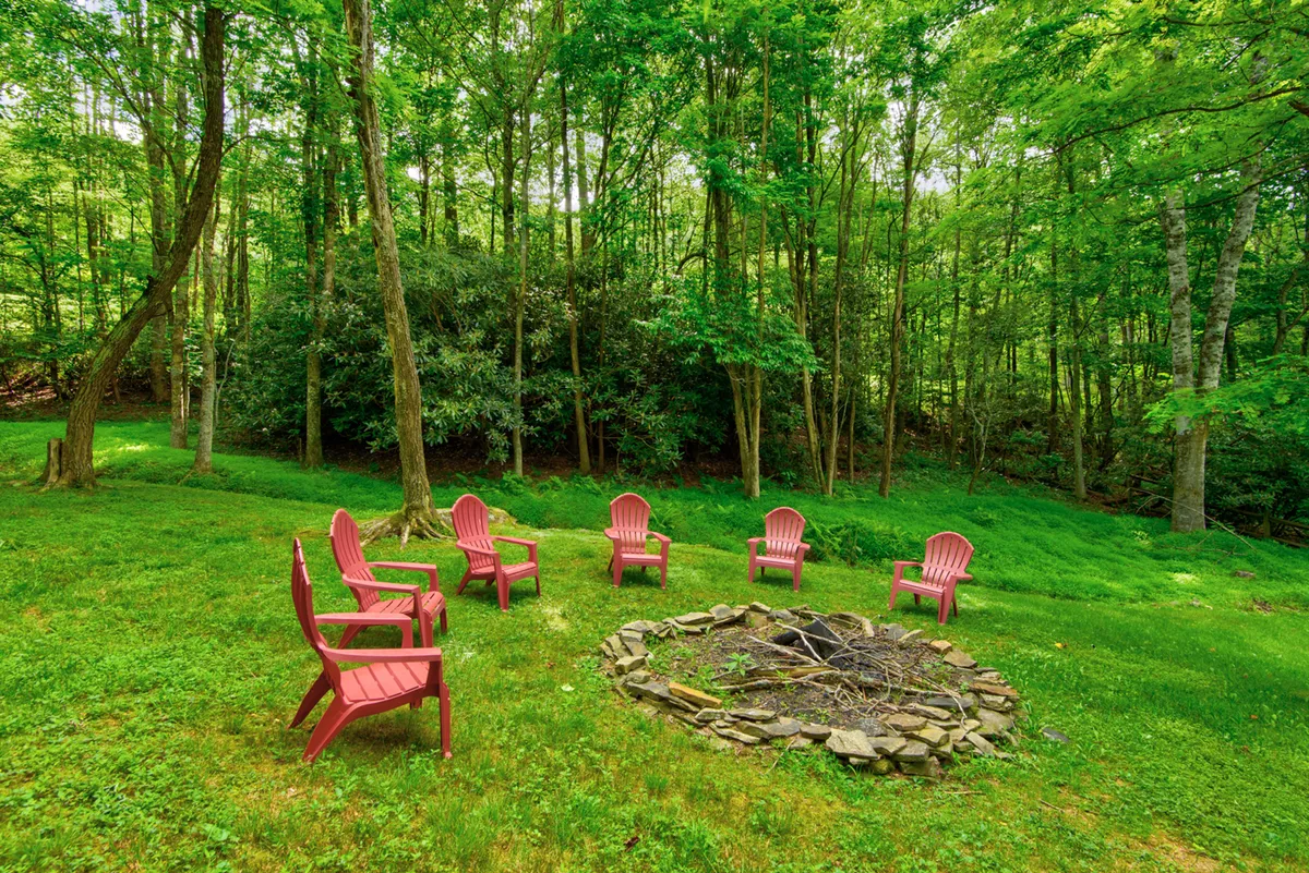 Red Adirondack chairs encircle a rustic stone fire pit in a grassy backyard framed by leafy trees. The scene radiates warmth and invites guests to gather for stories, s’mores, or simple relaxation under open skies.