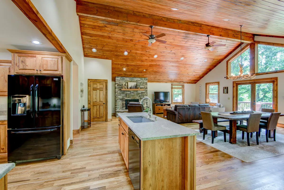 This angle highlights the open-concept kitchen, living, and dining area. The kitchen features a granite-topped island, wooden beams, and modern appliances, seamlessly flowing into the adjacent dining and living spaces with a stone fireplace and large windows.