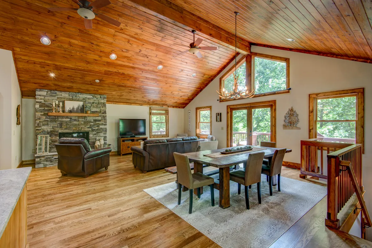 An inviting view of the living and dining area, featuring a stone fireplace, wooden ceiling, large windows, and a generous dining table. The warm lighting and arrangement create a welcoming atmosphere ideal for family dinners, game nights, or simply relaxing by the fire.