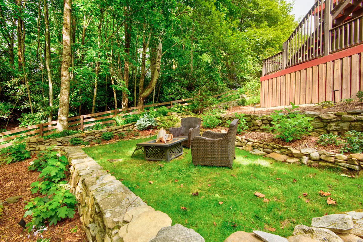 A lush backyard scene with an inviting outdoor seating area, a central fire pit, stone wall details, and vibrant greenery all around. The atmosphere is one of relaxation, socializing, and family fun, with space for kids and pets to play safely under supervision.