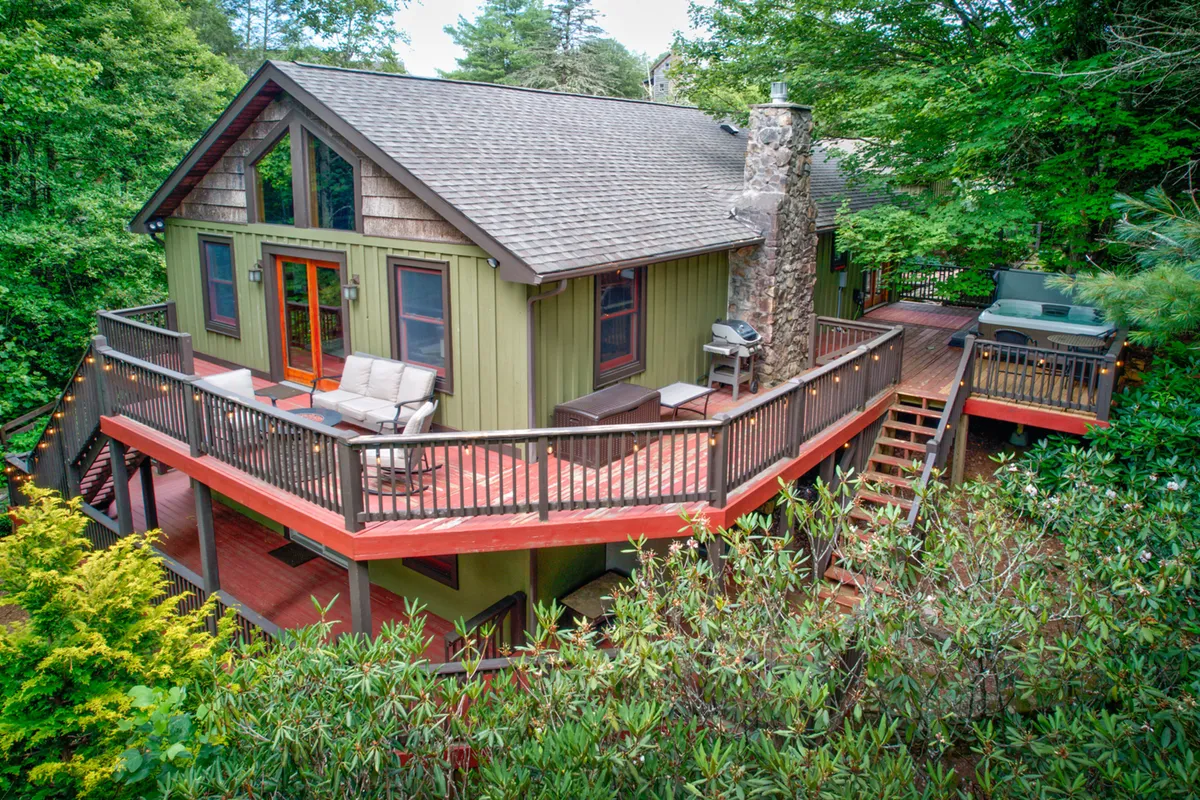 This image presents a spacious wooden deck adjoining the home, bordered by lush greenery and featuring large windows and a prominent stone chimney. Outdoor lounge chairs are visible, capturing the sense of relaxation and an inviting setup for gatherings or morning coffee.
